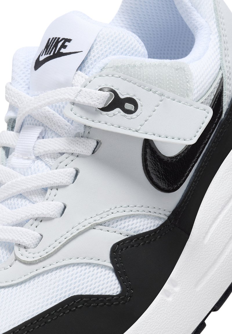 Witte sportschoen met zwarte accenten, synthetisch mesh bovenwerk, gevoerde tong en een band met een circulair logo. Zwarte Nike swoosh detail aanwezig.