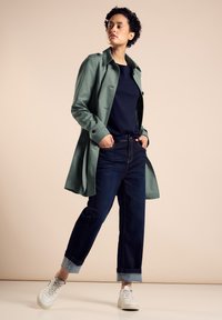 Ljusgrön trenchcoat över en marinblå topp, kombinerad med mörkblå jeans uppvikta vid fållarna och vita sneakers. Enkla, rena linjer och färger.