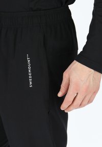 Czarne spodnie sportowe z teksturowanym wykończeniem, z elastycznym paskiem w talii i boczną kieszenią. Logo "SWEDEMOUNT" w kolorze białym na nogawce.