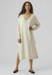 Robe tricotée crème, longueur genou, avec un décolleté en V profond, des manches longues, des fentes sur les côtés et un ourlet côtelé. Texture douce et coupe décontractée.
