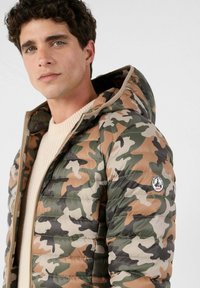 Gewatteerd jack met capuchon in een camouflagepatroon met tinten groen, bruin en beige. Heeft een doorgestikte textuur en een logopatch op de mouw.