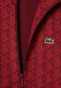 Veste rouge à fermeture éclair avec motif géométrique et petit logo de crocodile vert sur la poitrine, partiellement dézippée montrant une couche intérieure rouge unie.