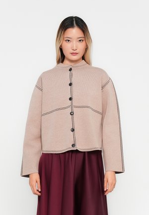 Junge Frau mit glattem Ombre-Haar, die eine beigen cardigan mit Knöpfen und schwarzen Stickdetails sowie einen fließenden burgunderfarbenen Rock trägt.