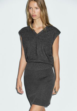 Woman wearing a dark gray sleeveless knit top and matching fitted skirt, with loose hair, standing against a plain light background.
