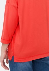 Rød, løs pasform top med trekvartærmer. Har en afrundet kant og sideslids. Stoffet ser glat og let ud.