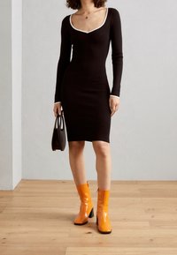 Robe noire à manches longues avec un col en V blanc, assortie à des bottes chevilles orange brillantes. Tient un petit sac à main noir. Fond en parquet.