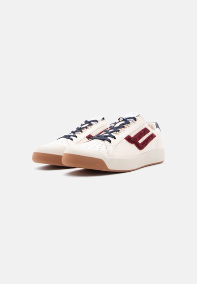 Ζευγάρι χαμηλά κρεμ αθλητικά παπούτσια με σκουρό μπλε κορδόνια, καφέ λαστιχένιες σόλες και κόκκινο ανάγλυφο λογότυπο στο πλάι, πάνω σε λευκό φόντο.