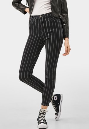 Persona con jeans ajustados negros con rayas verticales blancas, combinados con zapatillas Converse negras de caña alta y una chaqueta de cuero negra.