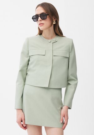 Vrouw met een lichtgroene cropped jas en bijpassende rok, grote donkere zonnebril en gouden opvallende oorbellen tegen een witte achtergrond.