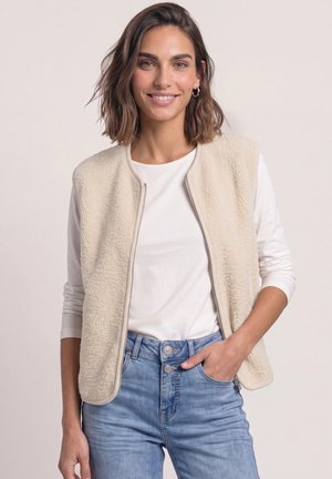 Femme aux cheveux bruns mi-longs portant une veste polaire crème sur un haut blanc à manches longues et un jean bleu clair, une main dans la poche.