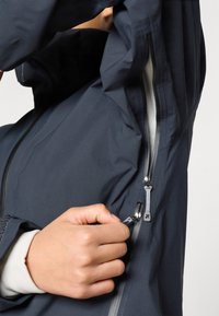 Tmavomodrá nepremokavá bunda s hladkým povrchom, vybavená zipsom a nastaviteľným sťahovacím šnúrkou. Detail ruky ťahajúcej zips.