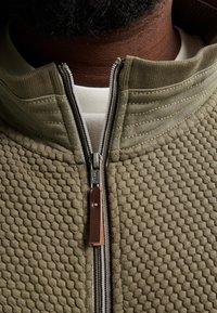 Olivgrön zip-jacka med texturerat bikake-mönster, som har en läderdetalj på dragkedjan och en hög krage.