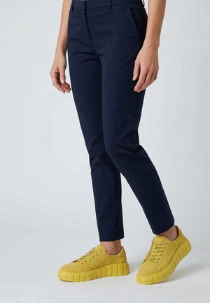 Frau trägt marineblaue Slim-Fit-Hose und leuchtend gelbe Plateausneaker und steht auf einem hellgrauen Boden.