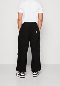 Schwarze Cargo-Hose mit lockerem Sitz, seitlichen Taschen und einem kleinen Logopatch auf der Rückseite. Kombiniert mit schwarzen und weißen Turnschuhen.