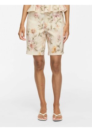 Shorts beiges doux à motif floral jusqu’aux genoux portés par une personne debout droite avec des sandales blanches sur un fond uni.