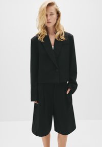 Čierny skrátený blazer s jedným gombíkom, skombinovaný s plisovanými čiernymi šortkami. Hladká textúra, minimalistický dizajn a prispôsobený strih.