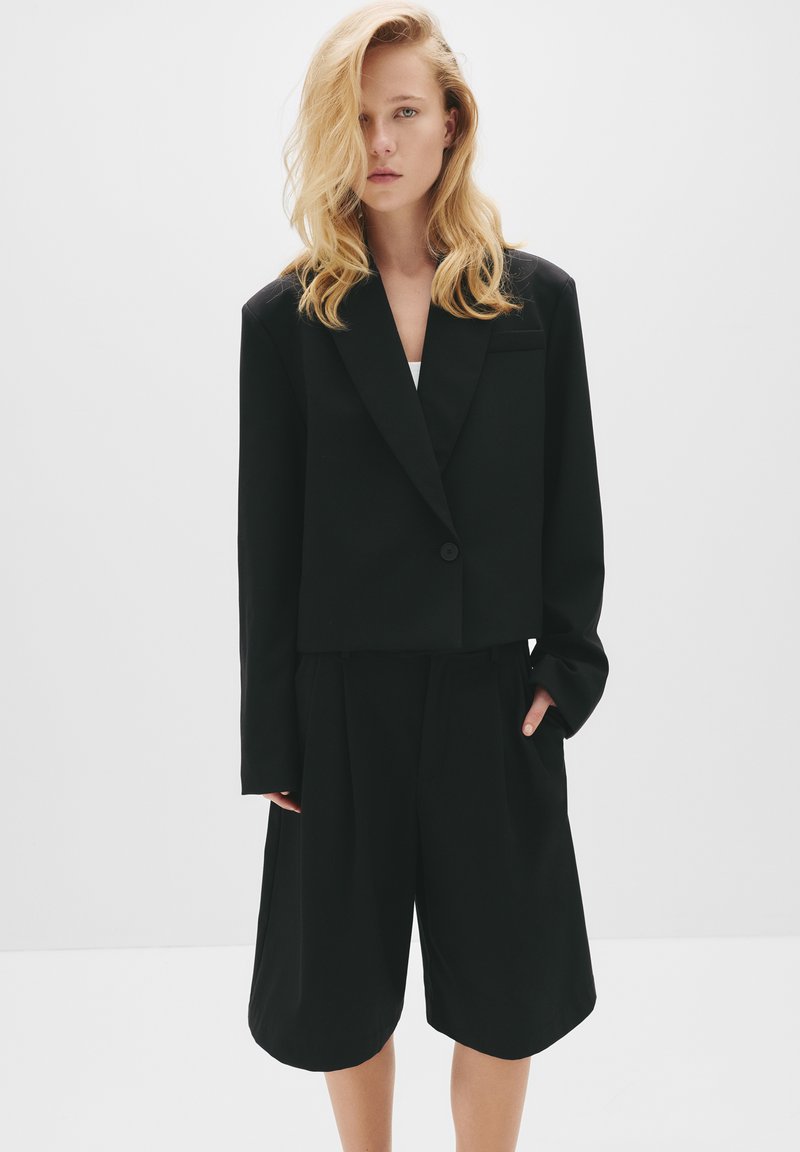 Čierny skrátený blazer s jedným gombíkom, skombinovaný s plisovanými čiernymi šortkami. Hladká textúra, minimalistický dizajn a prispôsobený strih.