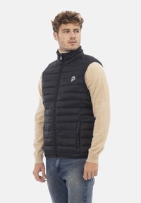 Schwarze Steppweste mit horizontaler Steppung, hohem Kragen, Reißverschluss und zwei Seitentaschen. Getragen über einem beigen Langarmshirt.