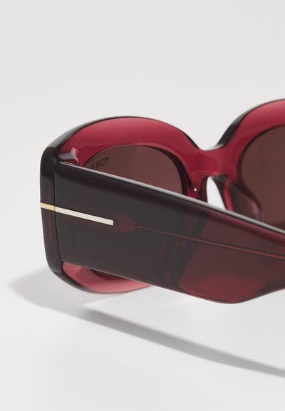 Óculos de sol com lentes vermelhas e um design robusto e oversized. Apresentam uma armação de plástico suave e um fino destaque em metal na haste.
