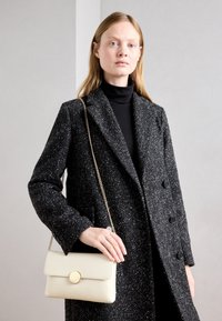 Manteau en laine texturée noir et blanc avec double boutonnage, porté sur un col roulé noir. Sac à main rectangulaire beige avec ferrures dorées.