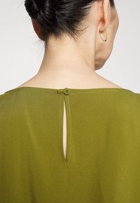 Blouse verte olive avec un détail de dos en trou de serrure, présentant un col rond et une petite fermeture à bouton à la nuque. Tissu lisse et léger.