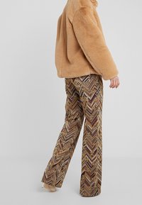 Tan nepleren jas gecombineerd met gemusterde broek met wijde pijpen die een zigzagpatroon vertonen in warme tinten van bruin, goud en bordeaux.