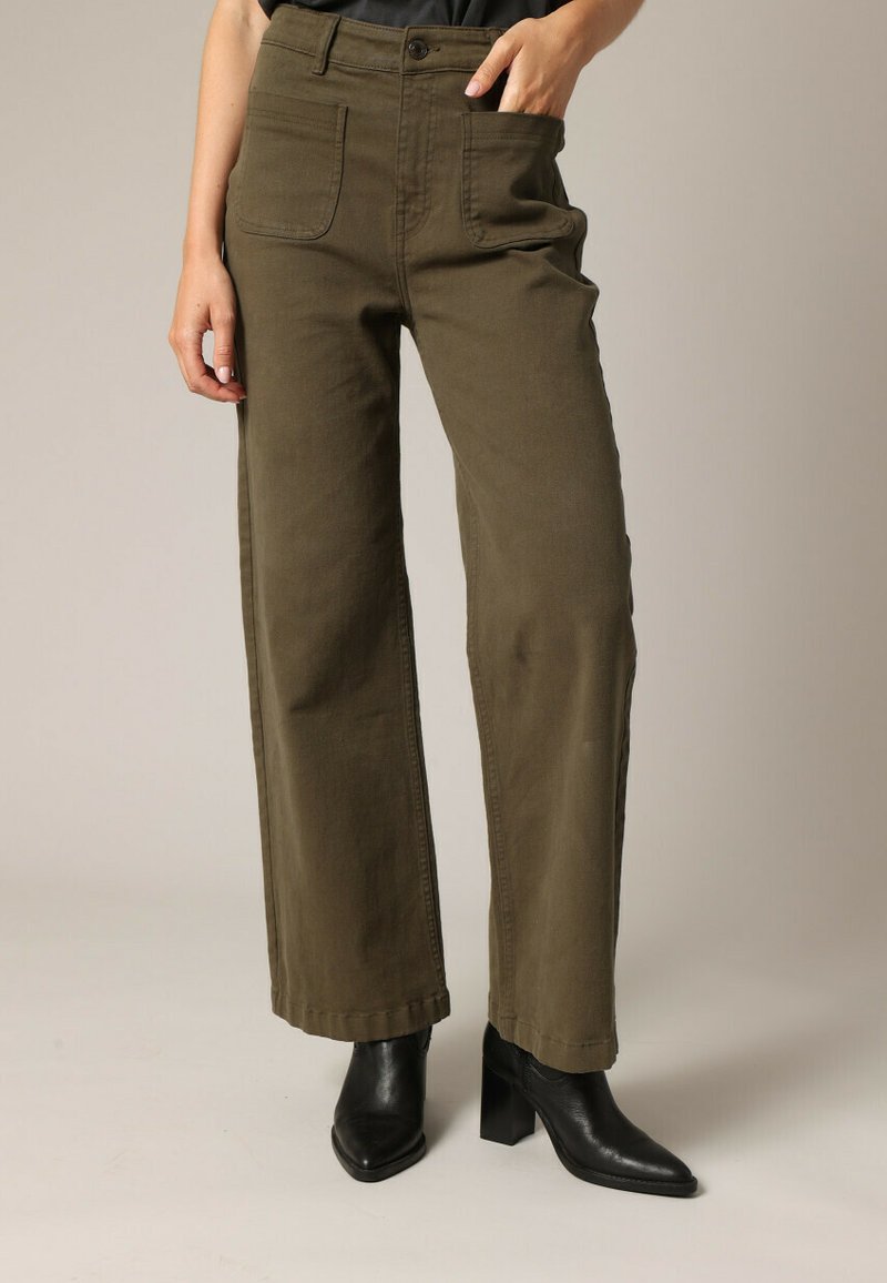 Pantalons larges olive en coton, dotés de poches frontales, d'une fermeture à bouton et d'accents cousus. Portés avec des bottes à talons noires.