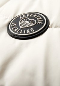 Rund sort og hvid lap på cremefarvet stof med hjerteemblem og teksten "ADVENTURE IS CALLING" omkring en solgrafik.