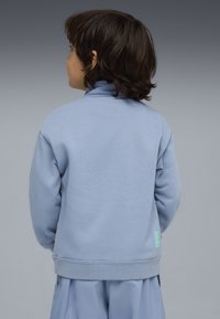 Bambino con capelli castani di lunghezza media, che indossa una felpa azzurra chiara e pantaloni coordinati, di spalle su uno sfondo grigio semplice.
