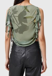 Femme portant un blouse à fleurs vertes avec des épaules dénudées et des détails drapés dans le dos, montrée de dos.