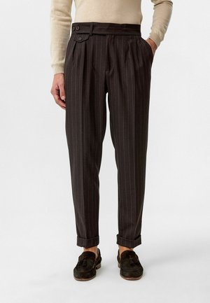 Bruine pinstriped broek met zijplooien, dubbele knooptaille en opgerolde manchetten, gemaakt van textuurstof. Gecombineerd met donkere instap schoenen.