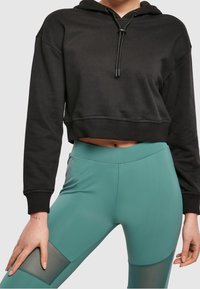 Felpa nera corta con cappuccio e coulisse, abbinata a leggings turchesi con dettagli in rete e una texture liscia.