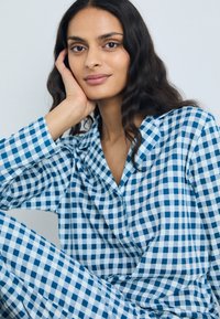 Conjunto de pijama em xadrez azul claro e branco, feito de tecido suave, com uma parte de cima com colarinho e calças combinando, usado por uma modelo de cabelo longo.