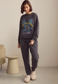 Šedá oversized mikina s grafickým potiskem a odpovídajícími joggery. Model nosí béžové chlupaté pantofle, čímž předvádí uvolněný, ležérní outfit.