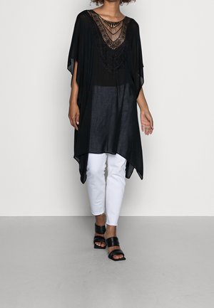 Femme portant une tunique noire ample avec détail en dentelle, pantalon blanc et sandales noires à talon bloc et bouts ouverts, marchant sur un fond uni.