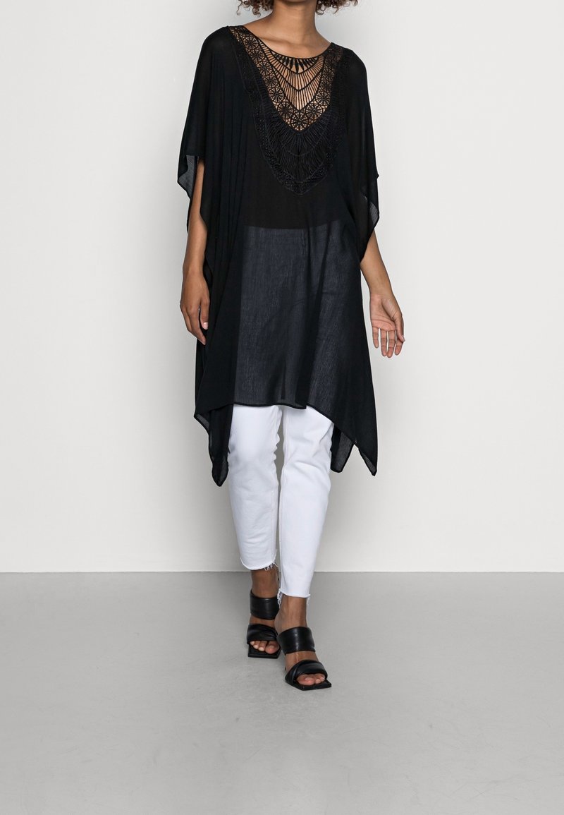 Femme portant une tunique noire ample avec détail en dentelle, pantalon blanc et sandales noires à talon bloc et bouts ouverts, marchant sur un fond uni.