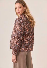 Mujer con cabello largo y ondulado de color rubio, vistiendo una chaqueta acolchada de estampado de hojas en tonos rosa y marrón, y una falda beige, mirando por encima del hombro.