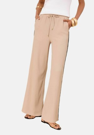 Woman wearing beige wide-leg trousers with black side stripes, a white sleeveless top, gold bangles, and black sandals with gold accents.