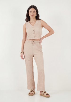 Donna in piedi che indossa un crop top beige testurizzato senza maniche con bottoni e pantaloni abbinati con coulisse, insieme a sandali beige.
