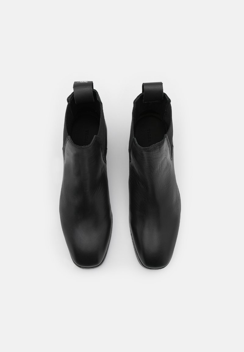 Para czarnych skórzanych botków typu slip-on za kostkę z kwadratowymi noskami i uchwytami, prezentowanych z góry na białym tle.