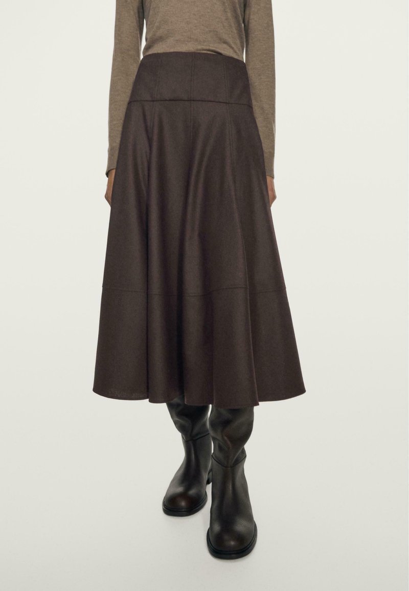 Jupe A-line marron à taille haute, en tissu texturé, avec des détails de coutures. Associée à des bottes en cuir noires montantes.