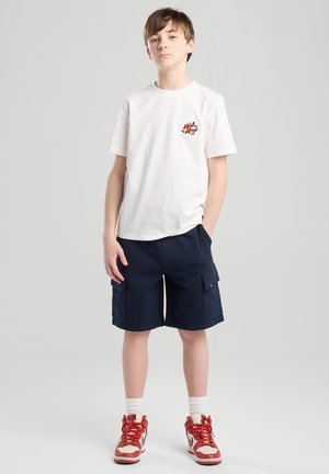 Weißes Baumwoll-T-Shirt mit kurzen Ärmeln und einem kleinen orangefarbenen Grafik. Navy Cargo-Shorts mit Taschen. Rote Sneaker mit weißen Akzenten.