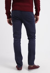 Mann trägt marineblaue Hose, burgunderfarbenen Pullover und passende burgunderfarbene Sneaker, steht mit entspannt neben dem Körper hängenden Armen.