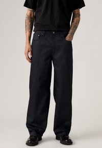 Jeans de pierna ancha color azul oscuro, hechos de denim, con un diseño clásico de cinco bolsillos, cierre frontal con botón y una textura suave. Se llevan calzado negro.