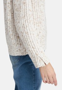 Textured cream sweater with beige speckles, ribbed cuffs, and a relaxed fit, paired with blue denim jeans.