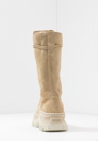 Bota de ante color marrón claro, de tiro alto, con una suela blanca gruesa y un tacón con marca. Presenta detalles de costura y una lengüeta en la parte superior para facilitar el uso.