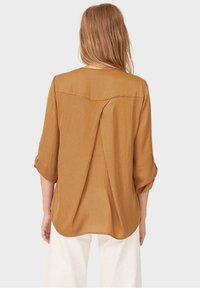 Camicia marrone leggera con pieghe sul retro e maniche arrotolate. Presenta un orlo arrotondato e una texture liscia.
