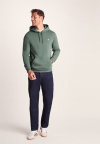 Smiling man stands with hands in pockets wearing a green hoodie, dark blue jeans, and white sneakers against a plain background.