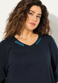 Laine paita tummansinisellä värillä, jossa on V-mallinen kaula-aukko, integroituja sinisiä yksityiskohtia ja hopeisia metalliosia hartioilla. Pehmeä kangas.