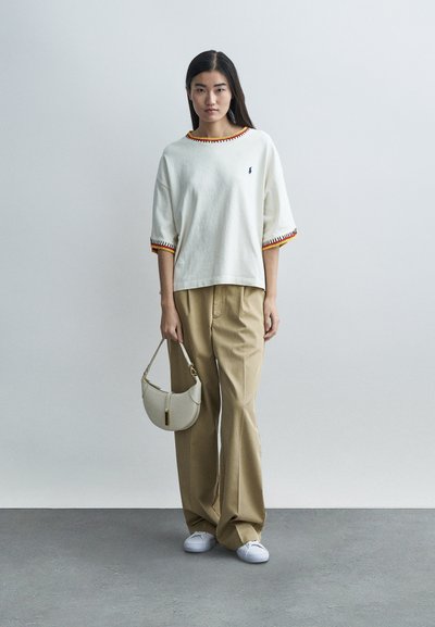 Biele oversized tričko s žíhanou textúrou a červeno-oranžovými akcentmi, skombinované s béžovými širokými nohavicami a svetlosivou kabelkou cez rameno.